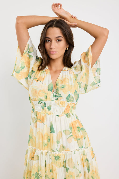 Woman wearing a honey-yellow floral maxi dress with flutter sleeves, v-neckline, and tiered flowing skirt, styled with neutral sandals.