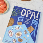 A cookbook titled 'OPA! Recipes for a Better Gathering' placed on a white marble countertop next to a charcuterie board with tomatoes, bread, olives, and cheese, with a plate of sliced tomato and cheese nearby.
