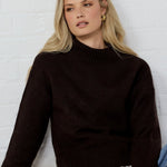 Woman wearing a dark brown sweater and blue jeans sitting against a white brick wall.
