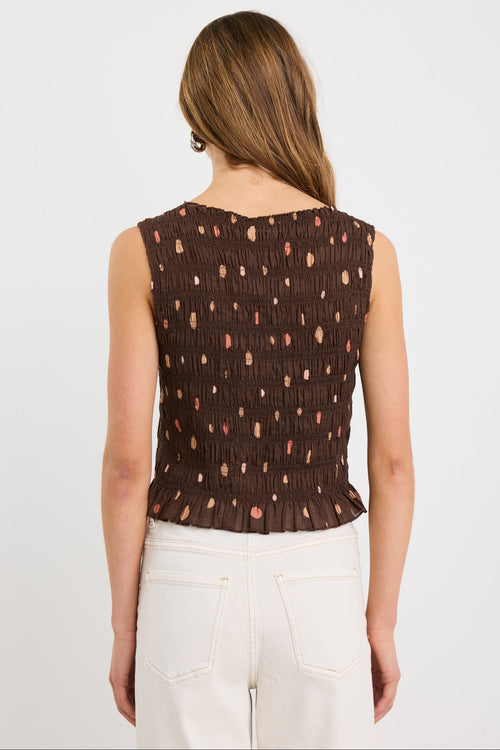 Woman wearing a sleeveless chocolate brown shirred top with scattered polka dot print and frilled hem, styled with ecru jeans.
