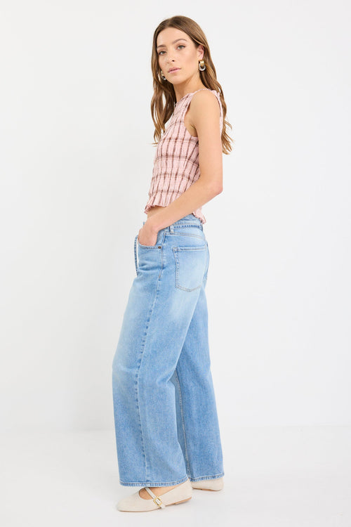 Woman wearing a blush pink sleeveless shirred top with check pattern and frilled hem, styled with light blue wide-leg jeans.