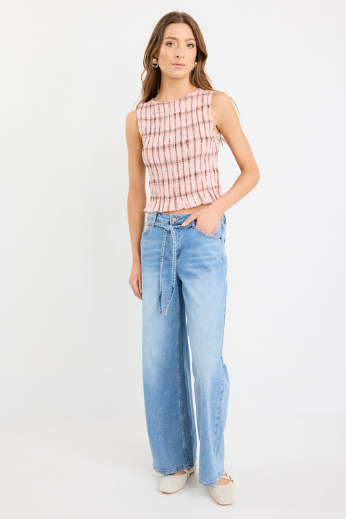 Woman wearing a blush pink sleeveless shirred top with check pattern and frilled hem, styled with light blue wide-leg jeans.