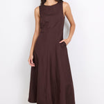 A woman modeling a sleeveless, dark brown midi dress with a high neckline and side pockets, paired with dark flat shoes, against a plain white studio background.