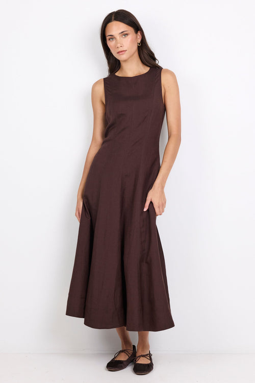 A woman modeling a sleeveless, high-neck, A-line midi dress in a deep chocolate brown color, standing against a plain white studio background.