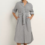 Woman wearing a black and white striped button-down midi shirt dress with a waist tie, rolled sleeves, and white sneakers.