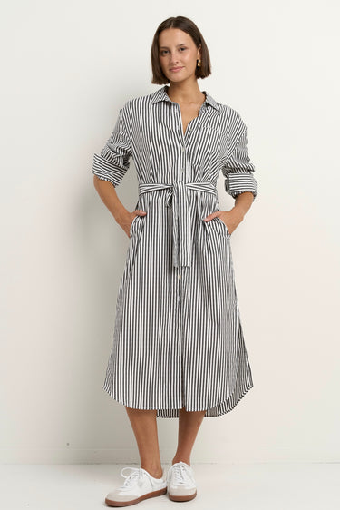 Woman wearing a black and white striped button-down midi shirt dress with a waist tie, rolled sleeves, and white sneakers.