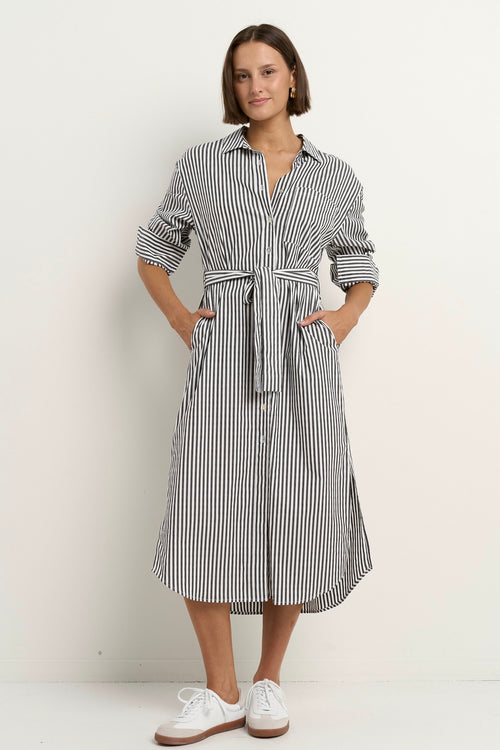 Woman wearing a black and white striped button-down midi shirt dress with a waist tie, rolled sleeves, and white sneakers.