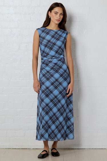 A woman stands against a white brick wall wearing a sleeveless, knee-length blue plaid dress with a gathered waist and black flats.