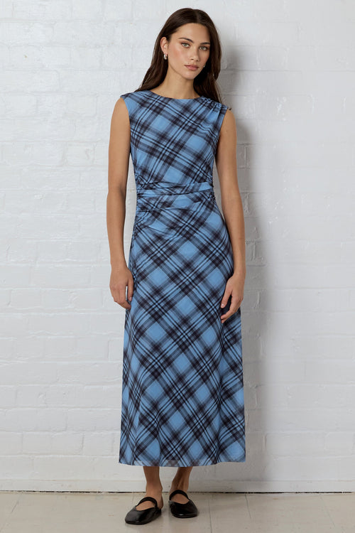 A woman stands against a white brick wall wearing a sleeveless, knee-length blue plaid dress with a gathered waist and black flats.