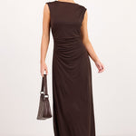 A woman modeling a sleeveless, floor-length dark brown dress with gathered ruching at the waist, styled with dark sunglasses, a matching handbag, and dark sandals against a plain white studio background.