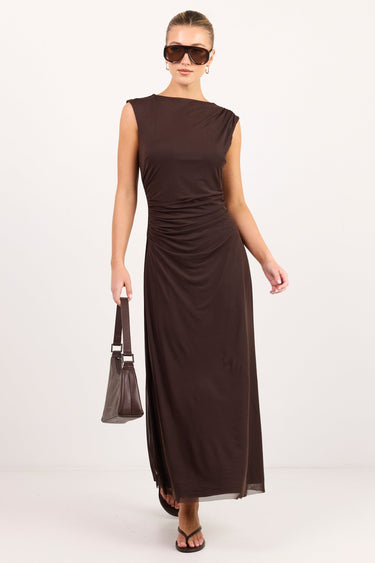 A woman modeling a sleeveless, floor-length dark brown dress with gathered ruching at the waist, styled with dark sunglasses, a matching handbag, and dark sandals against a plain white studio background.