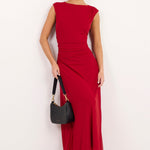 A woman modeling a sleeveless, off-the-shoulder red midi dress with a gathered waist, holding a black handbag against a plain white studio background.