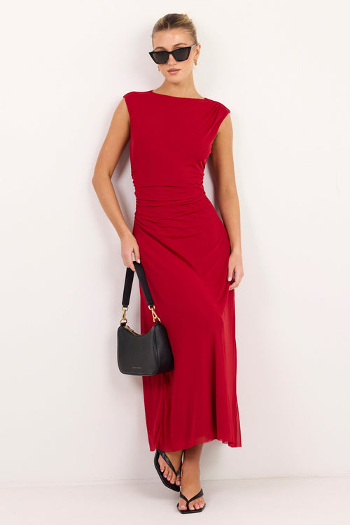 A woman modeling a sleeveless, off-the-shoulder red midi dress with a gathered waist, holding a black handbag against a plain white studio background.