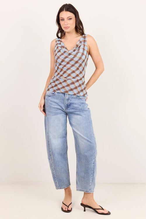A woman modeling a sleeveless cowl-neck top with a brown and beige checkered pattern, paired with light-wash wide-leg jeans and black heeled sandals, against a plain off-white studio background.