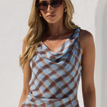 A woman modeling a sleeveless cowl-neck top with a brown and light blue plaid pattern, paired with white jeans, set against a bright white stucco wall with an archway.