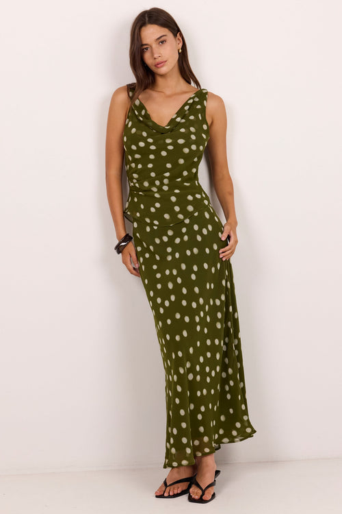 A woman modeling a sleeveless olive green maxi dress with a white polka dot pattern, standing against a plain white studio background.