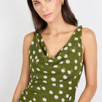 A woman wearing a sleeveless olive green cowl-neck top with a white polka dot pattern, paired with blue jeans, against a plain light gray background.