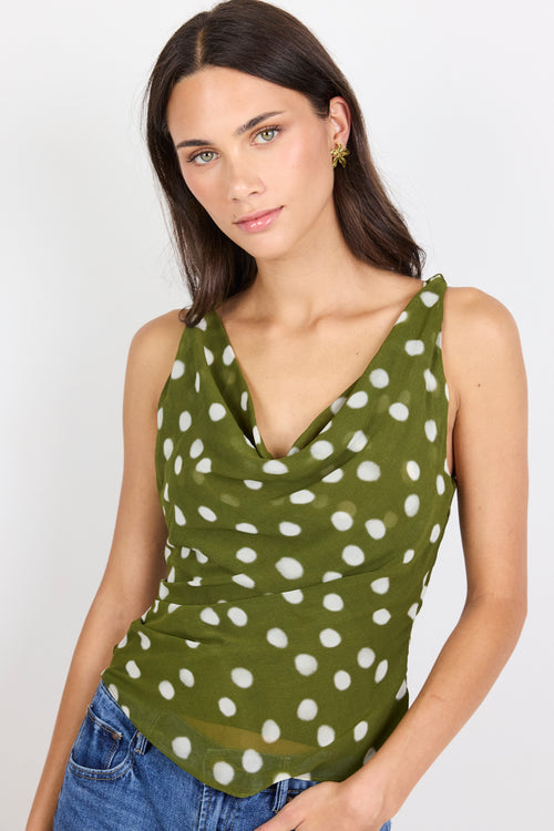 A woman wearing a sleeveless olive green cowl-neck top with a white polka dot pattern, paired with blue jeans, against a plain light gray background.
