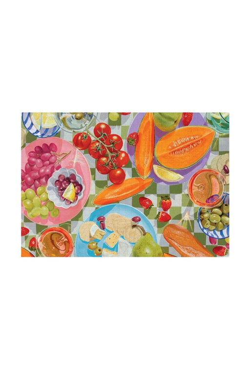 A rectangular placemat featuring a colorful, whimsical illustration of a picnic scene with plates of grapes, tomatoes, carrots, bread, cheese, and drinks on a green-and-white checkered blanket, shown against a plain white background.