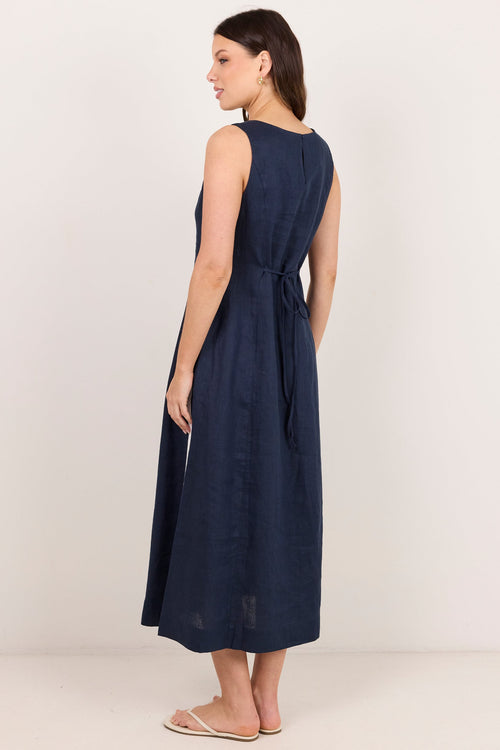 A woman modeling a sleeveless navy blue midi dress from the back, standing against a plain off-white studio background.