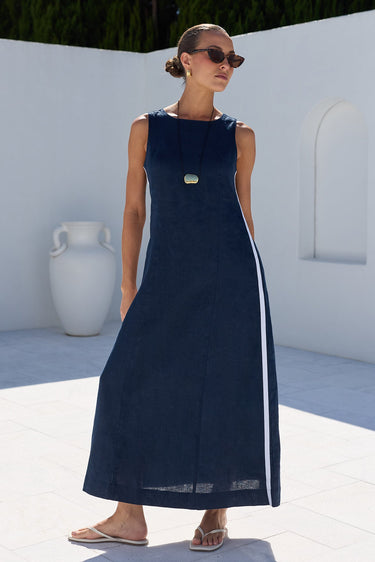 A woman modeling a sleeveless navy blue maxi dress with a white side stripe, standing in a bright, modern outdoor setting with white walls and dark green trees.