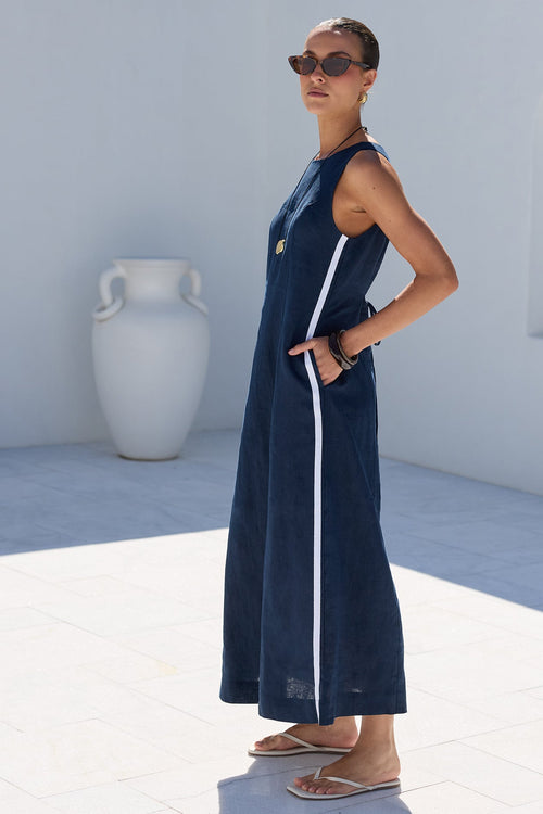 A woman modeling a sleeveless navy blue maxi dress with a white side stripe, standing in a bright white outdoor courtyard with dark green trees in the background.