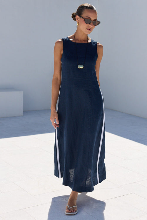 A woman wearing a sleeveless navy blue midi dress with white side stripes and a small gold chest emblem, standing in a bright white architectural courtyard.