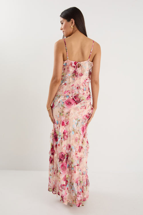 Woman wearing a strappy floral bias-cut maxi dress in pink and pastel tones with cascading ruffle layers, styled with neutral sandals.