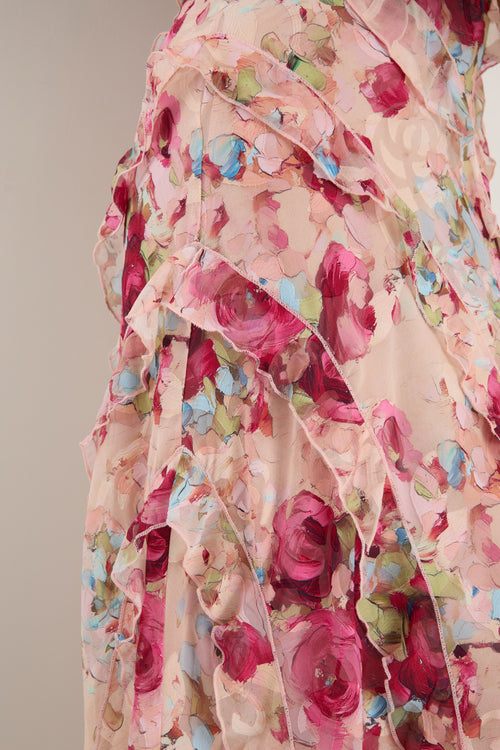 Woman wearing a strappy floral bias-cut maxi dress in pink and pastel tones with cascading ruffle layers, styled with neutral sandals.