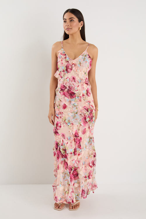 Woman wearing a strappy floral bias-cut maxi dress in pink and pastel tones with cascading ruffle layers, styled with neutral sandals.