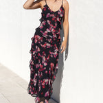 A woman wearing a black ruffled maxi dress with a pink and red floral print, standing against a white stucco wall next to a pool.