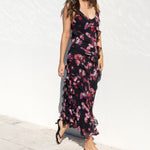 A woman wearing a sleeveless black maxi dress with a pink and red floral print, black sunglasses, and black sandals, standing outdoors against a white wall.