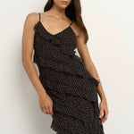 Woman wearing a black bias-cut polka dot dress with delicate straps and cascading ruffles, standing against a neutral wall.