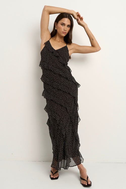 Woman wearing a black bias-cut polka dot dress with delicate straps and cascading ruffles, standing against a neutral wall.