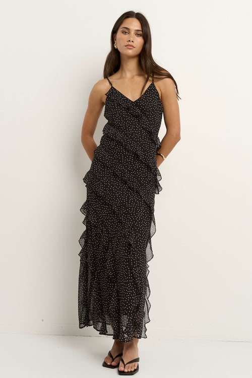 Woman wearing a black bias-cut polka dot dress with delicate straps and cascading ruffles, standing against a neutral wall.