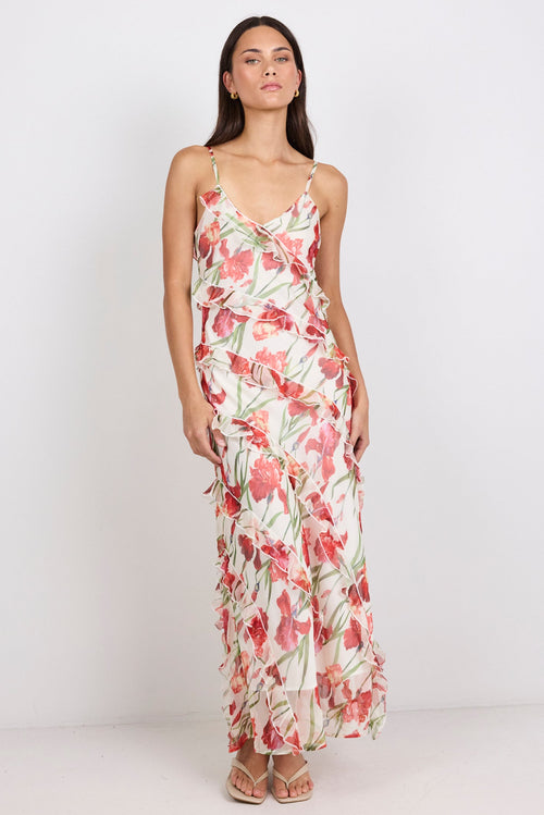 A woman modeling a sleeveless maxi dress with thin straps and a ruffled tiered skirt, featuring a pink and green tropical floral print on a cream background, set against a plain white studio backdrop.