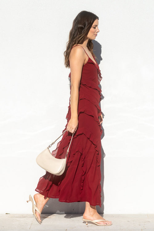 A woman in a long burgundy ruffled maxi dress walks in profile against a bright white wall, carrying a cream shoulder bag.