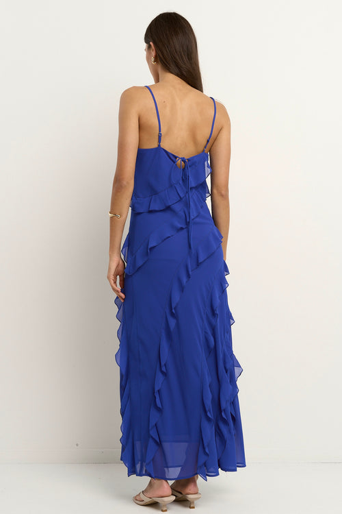 Woman wearing a bright electric blue bias-cut maxi dress with delicate straps and cascading ruffle details, standing against a neutral background.