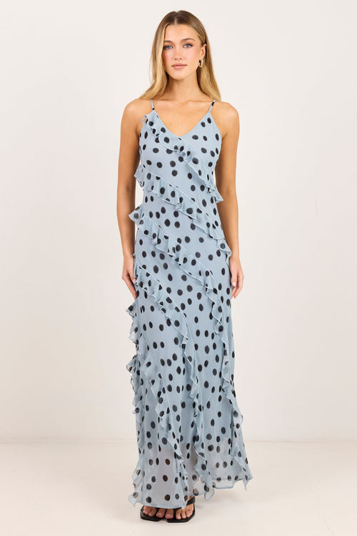 A woman modeling a light blue sleeveless maxi dress with black polka dots, photographed against a plain off-white studio background.