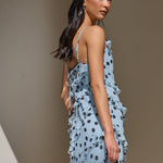 A woman wearing a light blue ruffled maxi dress with black polka dots, holding a black handbag, photographed in a warm-toned indoor setting.