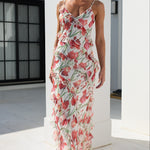 A woman models a sleeveless, ruffled maxi dress with a red and pink floral print on a white background, walking in a bright, modern white outdoor corridor.