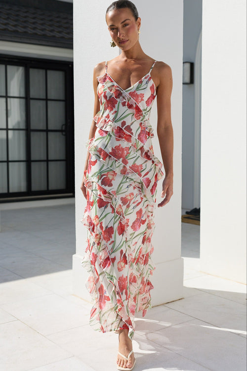 A woman models a sleeveless, ruffled maxi dress with a red and pink floral print on a white background, walking in a bright, modern white outdoor corridor.
