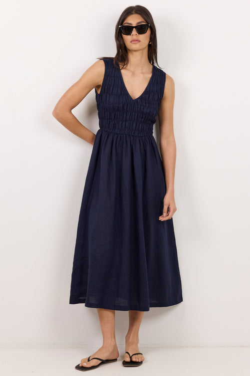 A woman modeling a sleeveless navy blue midi dress with a smocked bodice and V-neckline, paired with black sandals and sunglasses, standing against a plain white studio background.