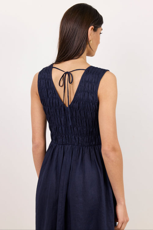 A woman shown from the back wearing a sleeveless dark navy dress with a deep V back and tie detail, photographed against a plain light background.