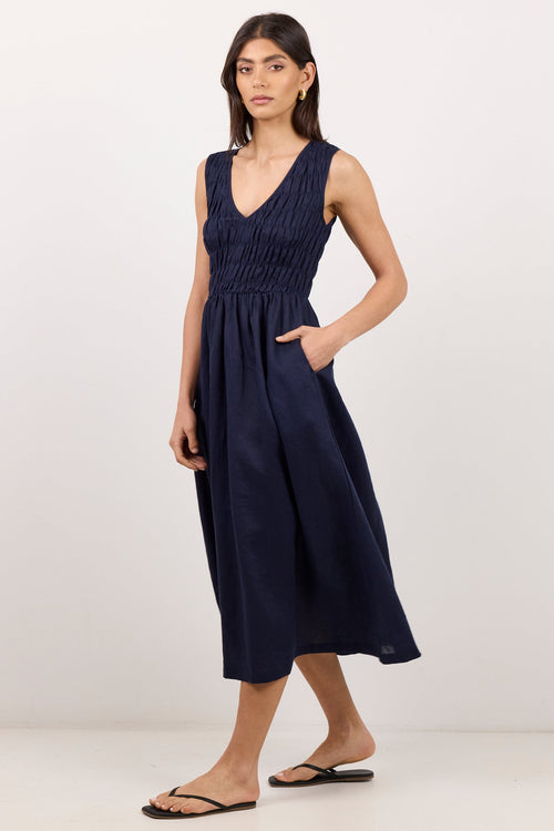 A woman modeling a sleeveless navy blue midi dress with a smocked bodice, V-neckline, and side pockets, paired with black flip-flop sandals against a plain white studio background.