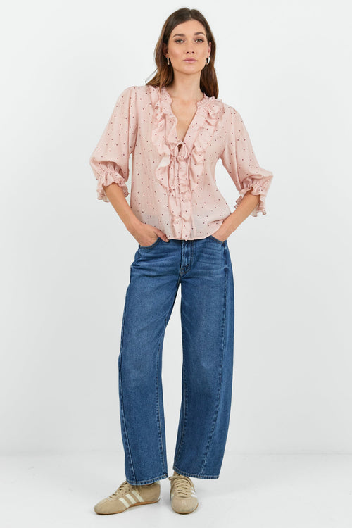 A woman modeling a light pink ruffled blouse with a subtle white pattern, paired with wide-leg blue jeans and beige sneakers, standing against a plain white studio background.