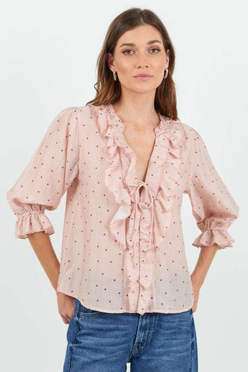 A woman modeling a light pink long-sleeve blouse with a delicate polka dot pattern, ruffled trim, and a tie-front detail, paired with blue jeans against a plain white studio background.