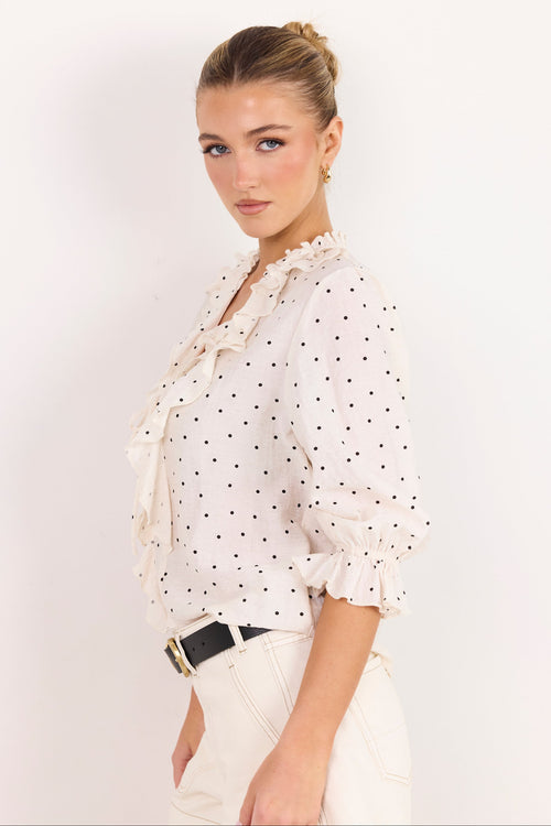 A woman modeling a white long-sleeve blouse with black polka dots, ruffled trim, and a tie-neck detail, paired with cream pants and a black belt, against a plain white background.