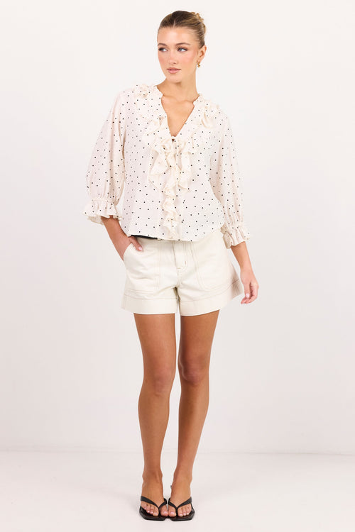 A woman modeling a white eyelet blouse with small black polka dots and a tie neckline, paired with white shorts and black sandals, standing against a plain white studio background.