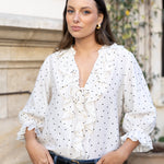 A woman wearing a white long-sleeve blouse with black polka dots and ruffle details, paired with blue jeans and a black belt, standing outdoors in front of a stone wall and greenery.